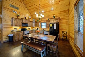 A wooden cabin interior with a dining table, benches, a chandelier, wooden cabinets, a microwave, and a refrigerator.