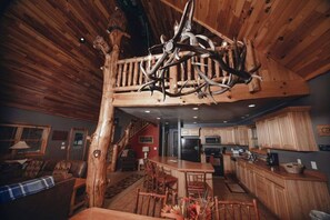 A rustic interior with a high ceiling, wooden beams, a dining area, kitchen, and a staircase.