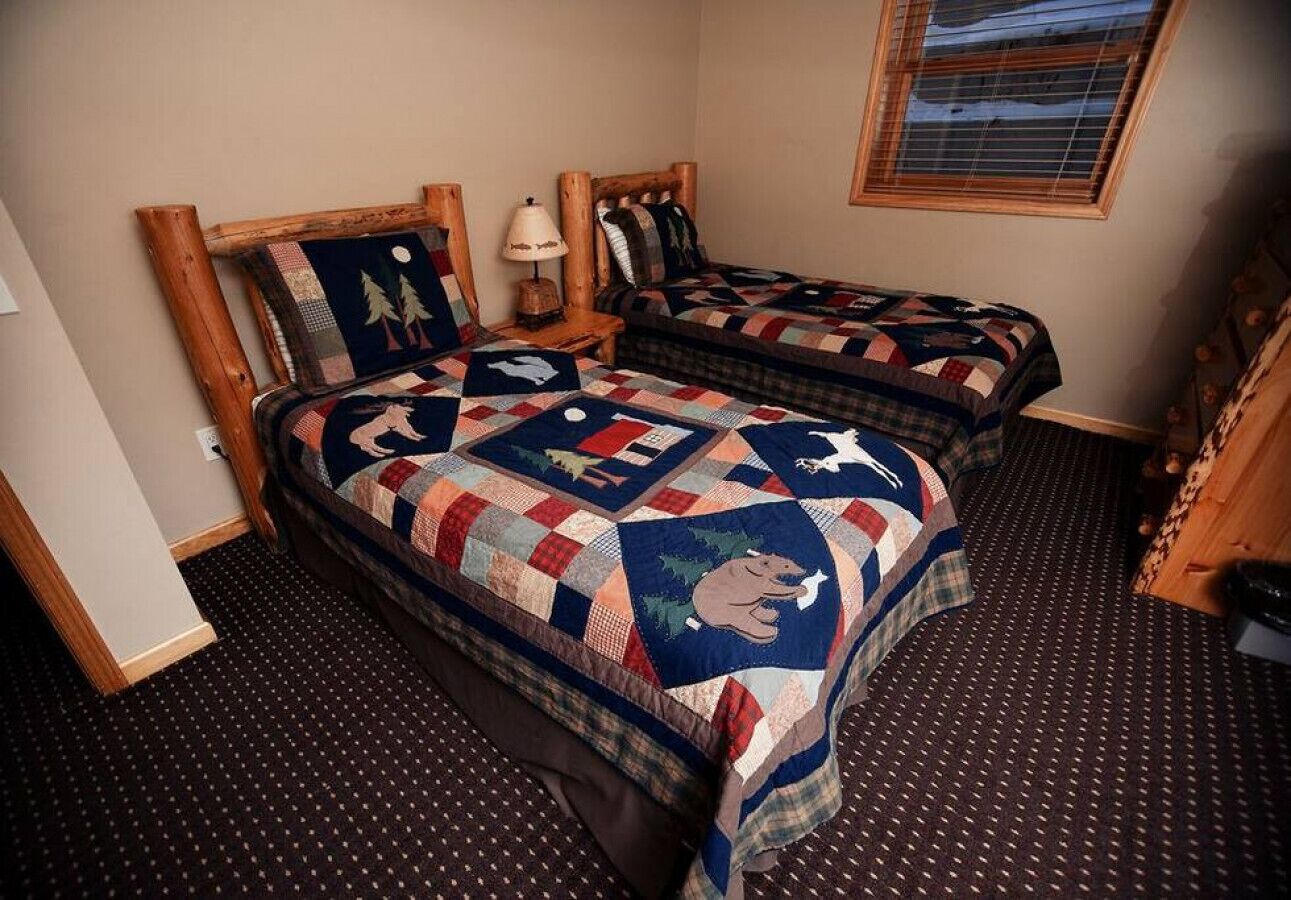 A room with two beds, each with a quilt featuring a bear design. There is a window with blinds, a lamp on the nightstand, and a door on the left.