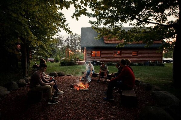 Um grupo de pessoas sentadas ao redor de uma fogueira em uma área arborizada, com uma cabana de toras ao fundo.