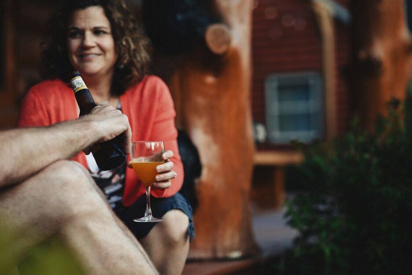 A person holding a bottle and a glass of orange liquid, seated outdoors with a wooden structure in the background.