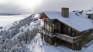 Un grand chalet en bois doté d’un balcon, entouré d’arbres couverts de neige et offrant une vue sur un paysage enneigé.