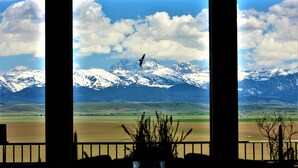 Une vue sur une chaîne de montagnes aux sommets enneigés, un ciel dégagé et, au premier plan, une zone délimitée par une clôture, où l’on aperçoit quelques plantes.