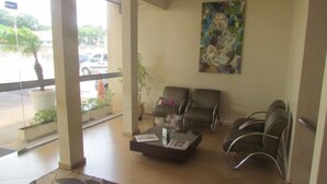 A reception area with a glass wall, two seating areas with chairs, a coffee table with a magazine, and a painting on the wall. - Hotel Capri (Medianeira)