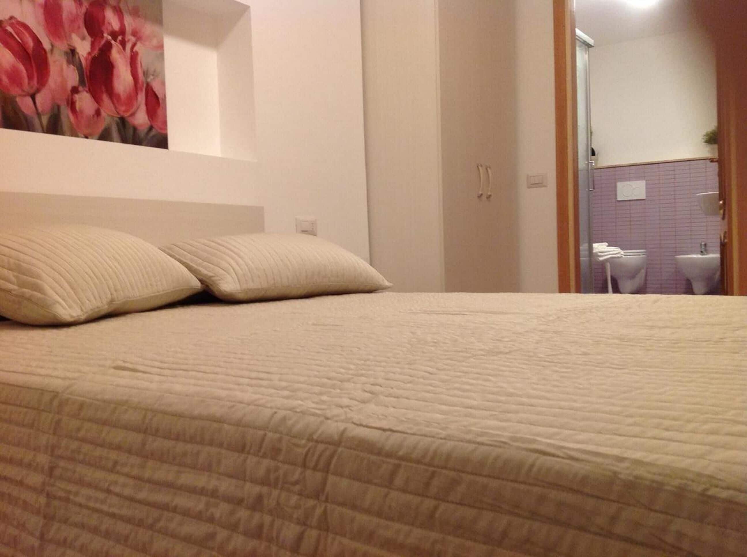 A bed with a beige quilt, two pillows, and a painting of red tulips on the wall.