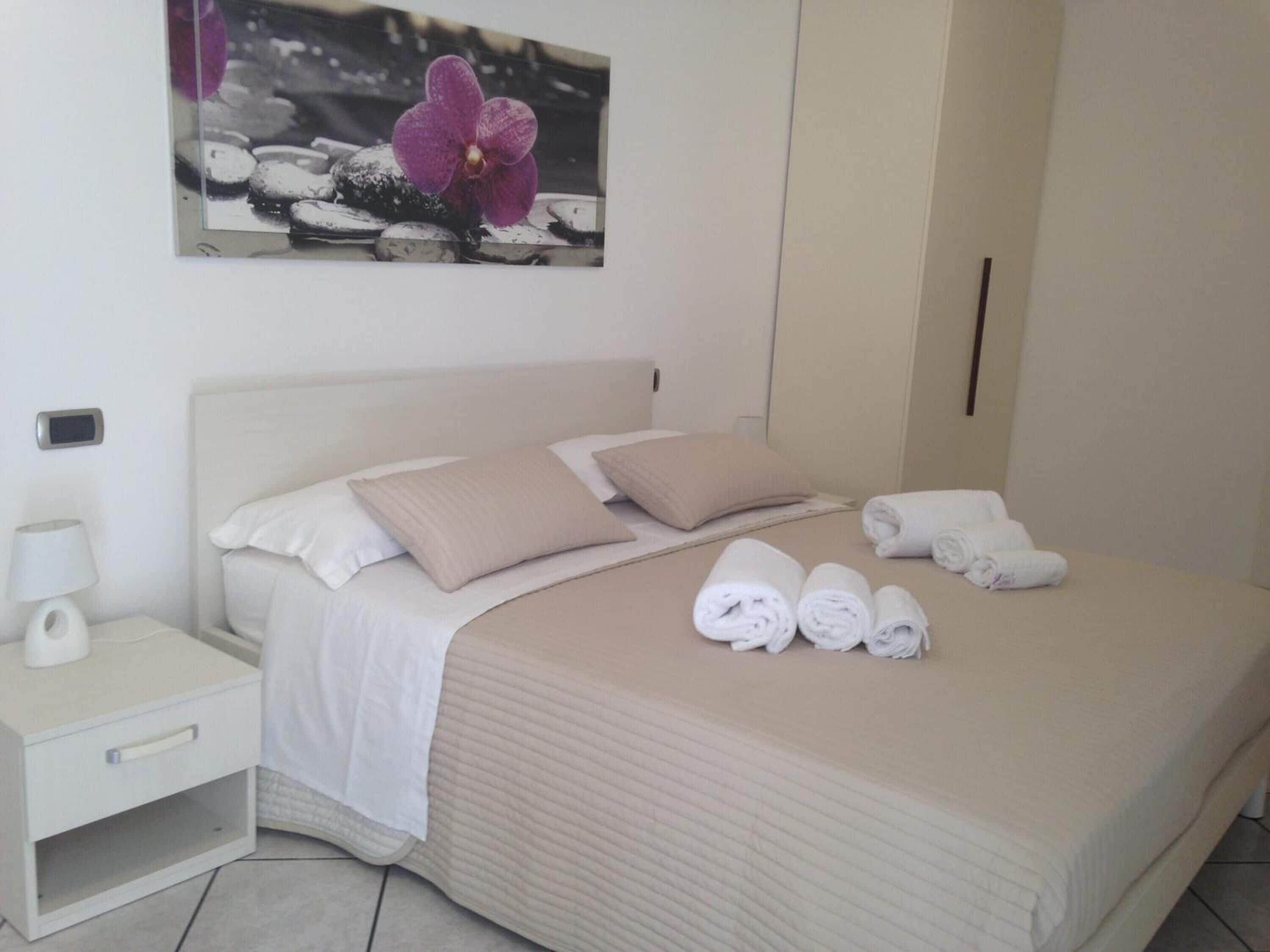Een net opgemaakt bed met witte lakens en beige kussens, een nachtkastje met een lamp, en een opgerolde handdoek op het bed.