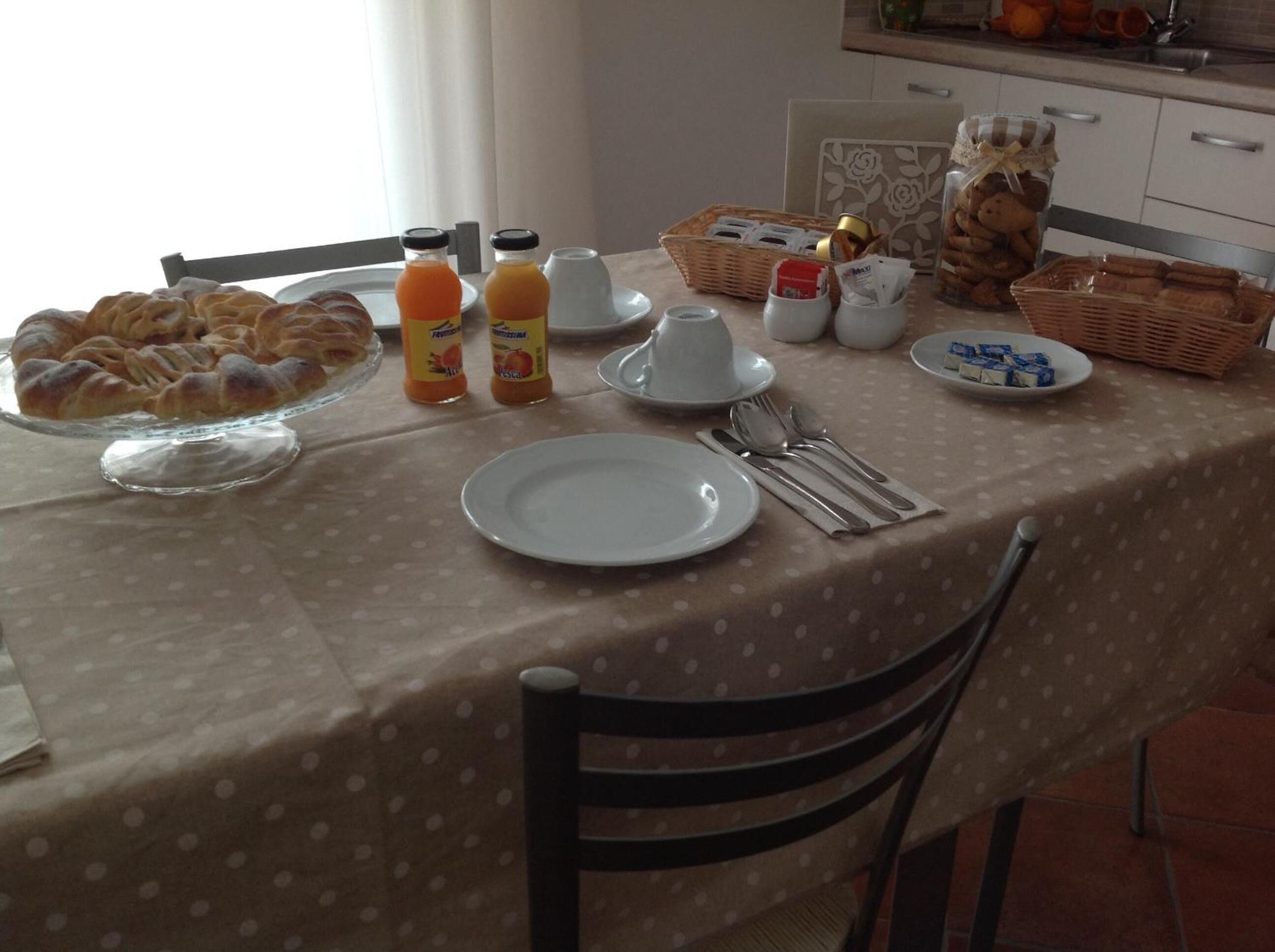 Una mesa de desayuno con mantel de lunares, un plato de pasteles, botellas de jugo, una cesta de pan y alimentos para el desayuno.