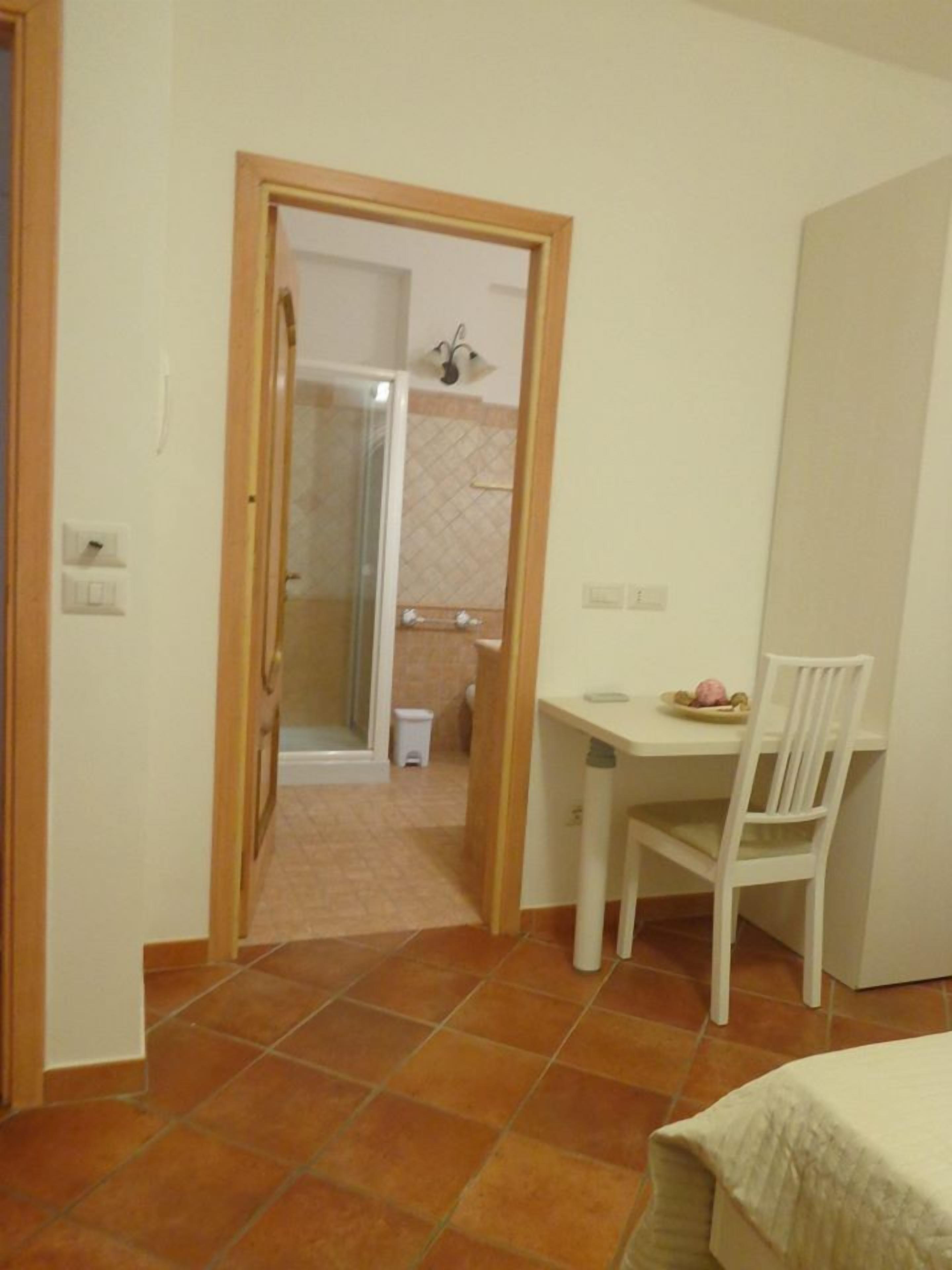 A small room with a tiled floor, a white chair, a table with a plate and a bowl, and a shower area visible through a doorway.