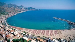 An aerial view of a coastal town with a beach, a pier, and buildings along the coastline. - A Casa Di Amici (Giardini Naxos)