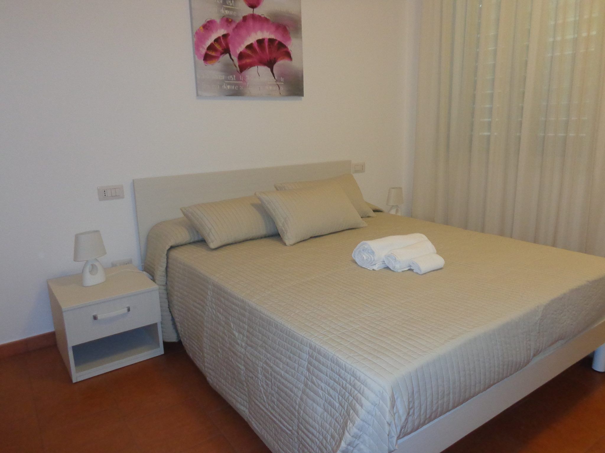 Ein ordentlich bezogenes Bett mit einer beige farbenen Tagesdecke und Kissen, ein weißer Nachttisch mit einer Lampe und ein an der Wand hängendes Kunstwerk über dem Bett.