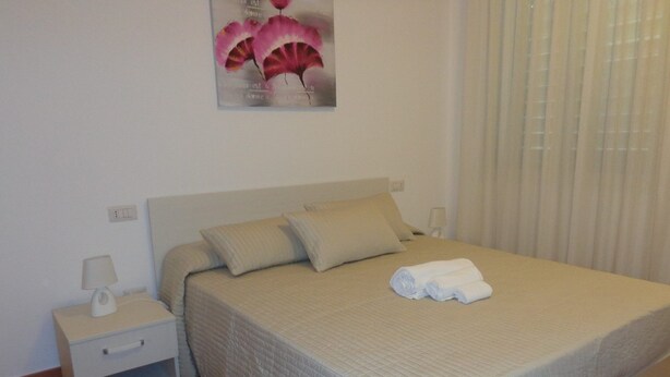 A neatly made bed with a beige bedspread and pillows, a white nightstand with a lamp, and a wall-mounted artwork above the bed. - A Casa Di Amici (Giardini Naxos)