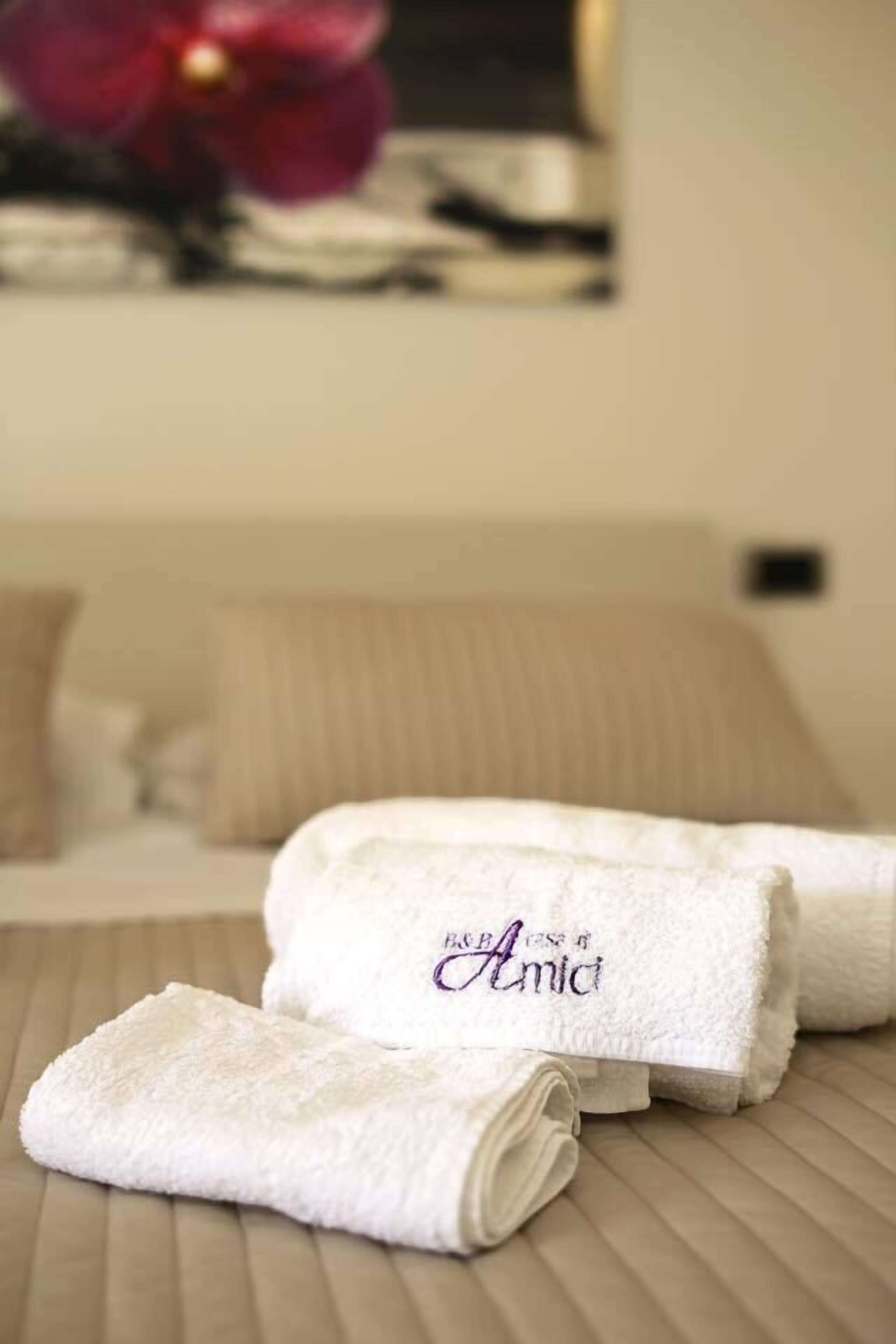 Een keurig gevouwen witte handdoek met de geborduurde tekst "B&B Amia", op een houten oppervlak in een kamer met een bed en een schilderij op de achtergrond.