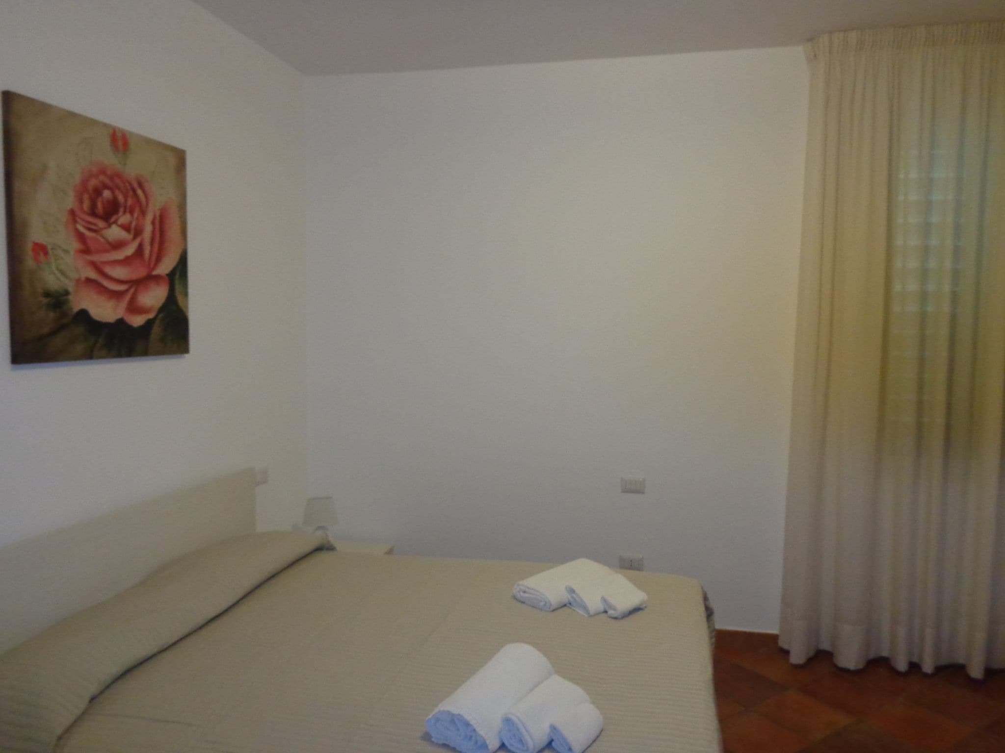 A hotel room with a bed, a painting of a rose, and a window with curtains.
