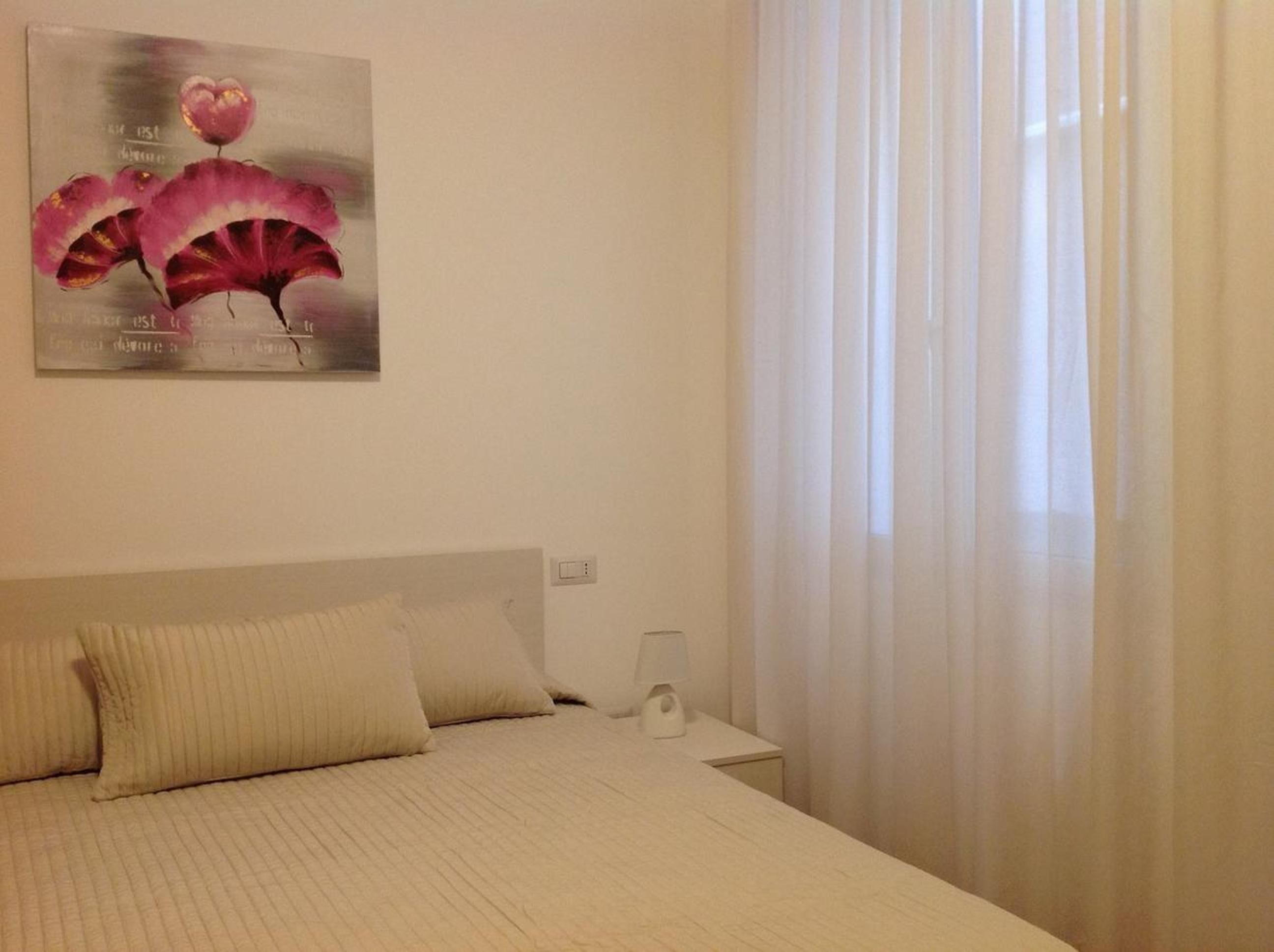 Un dormitorio con cama, mesita de noche, una obra de arte en la pared y cortinas translúcidas.