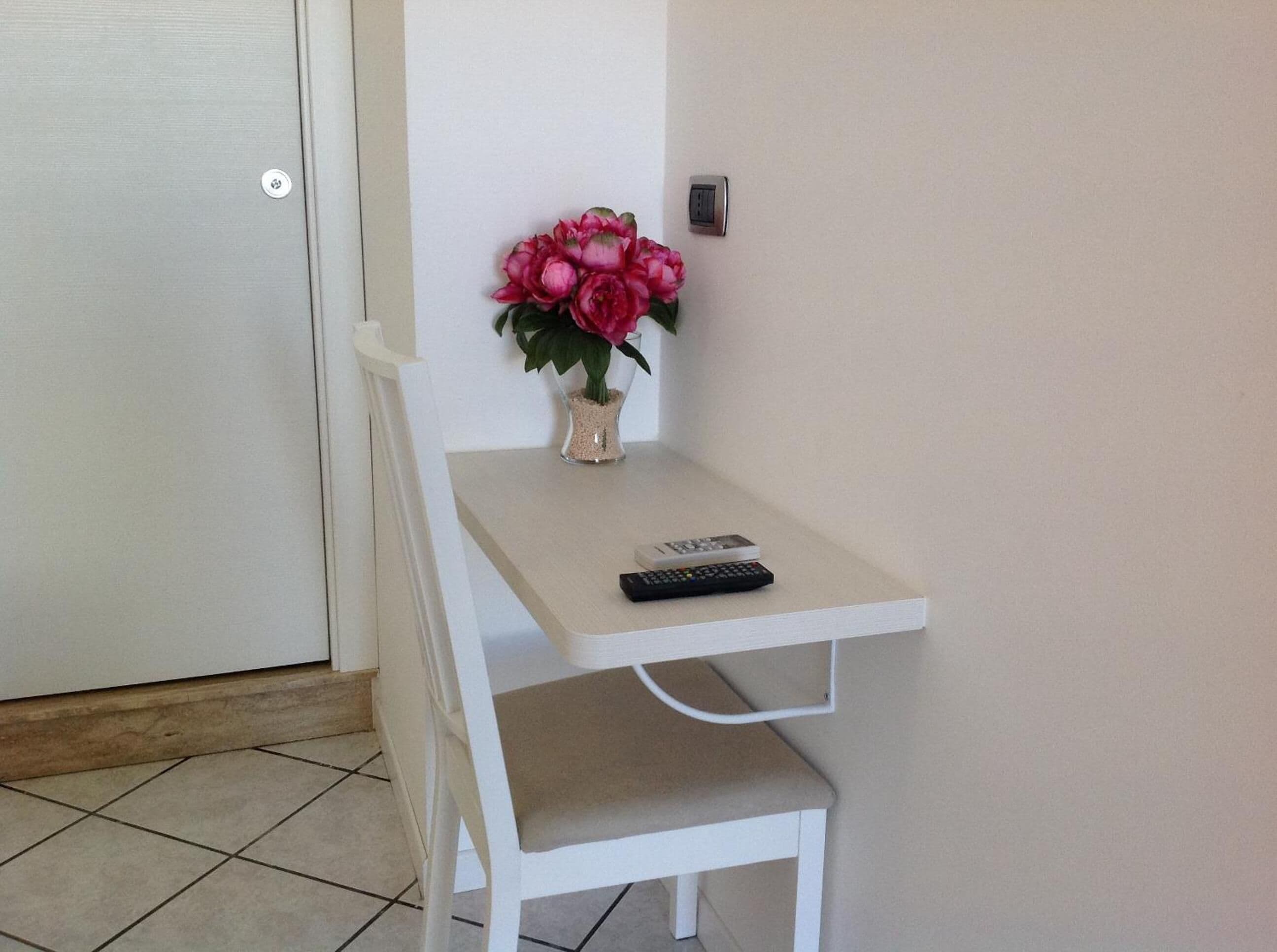A small white desk with a vase of pink flowers and two remote controls.