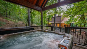 A hot tub with a wooden deck and metal railing, surrounded by trees and a cabin in the background.