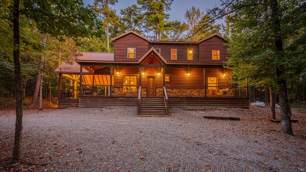 A rustic log cabin with a covered porch, multiple windows, and a gravel driveway surrounded by trees.