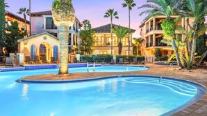 A swimming pool with a tiled edge, surrounded by palm trees and a building with a tiled roof. - Short Walk to Beach • Pool • Pets Allowed • (Miramar Beach)