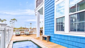 Un área de piscina con cubierta de madera, baranda blanca y un edificio azul al fondo.