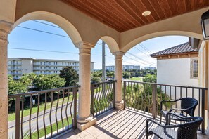 A balcony with a wooden ceiling, columns, and a railing. There are two wicker chairs.