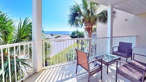 A balcony with wicker furniture and a glass table, overlooking a beach and palm trees.
