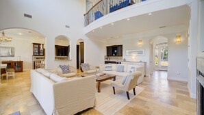 A spacious living room with a large sofa, a coffee table, and a fireplace. There is a kitchen in the background with a dining area visible through an archway.