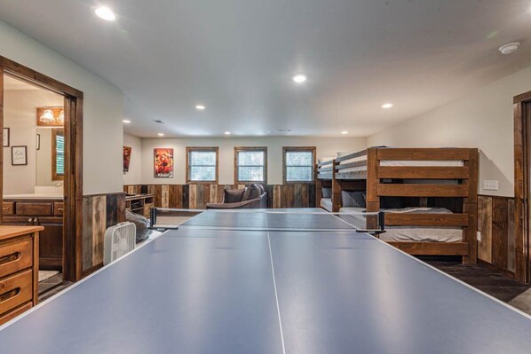 A room with a ping pong table, bunk beds, a sofa, and a fan.