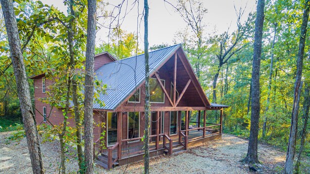 A cabin with a porch, surrounded by trees and a gravel driveway. - Hot Tub • Game Loft • Walk to Local Attractions • Sleeps 10 (Broken Bow)