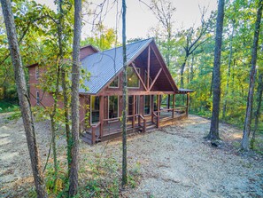 A cabin with a porch, surrounded by trees and a gravel driveway.