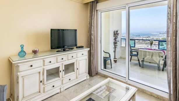 Un balcone con un tavolo e sedie, una televisione su un mobile, e vista sul mare e sul porto turistico.