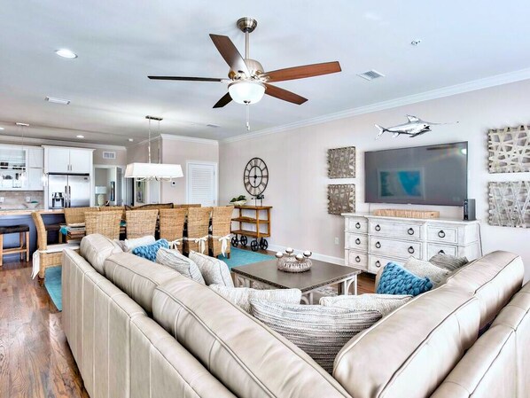 A modern living room with a beige leather sofa, a dining area with wicker furniture, a ceiling fan, and a flat-screen TV mounted on the wall. - Luxury Townhome, Golf Cart, Short Walk to Beach, Pool, Sleeps 13 (Miramar Beach)