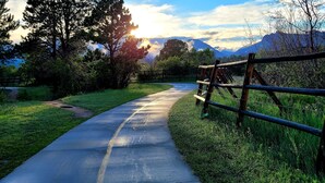 A paved pathway with a wooden fence on one side, surrounded by grass and trees, leading towards a mountainous landscape under a clear sky. - Charming 2 Bedroom w/ Private Hot Tub and Mountain views! (Estes Park)