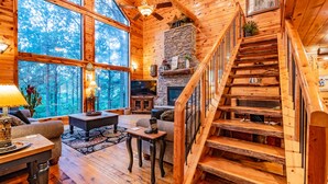 A cozy cabin interior with a stone fireplace, wooden staircase, and large windows offering a view of the forest.