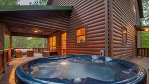 A wooden cabin with a hot tub on a wooden deck, a covered patio, and a view of the surrounding forest.