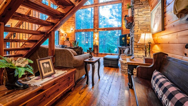 A cozy cabin interior with a wooden staircase, large windows, a fireplace, and a comfortable seating area.