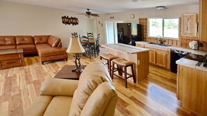 Amplia sala de estar con un sofá de cuero marrón, un sillón color beige y una mesa de comedor de madera con sillas. Se ve una cocina en el fondo con armarios de madera y un refrigerador.