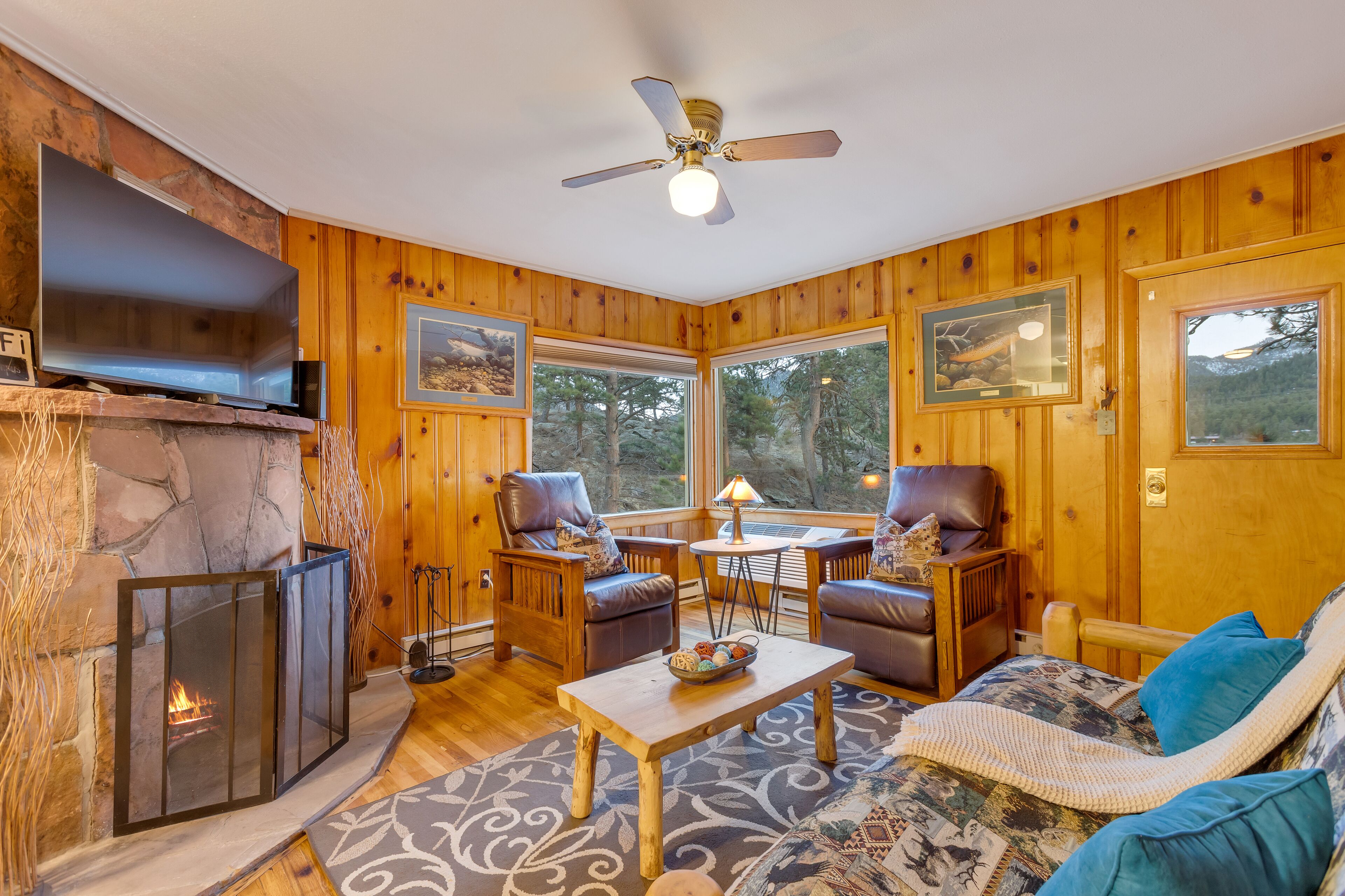 A cozy cabin interior with a fireplace, wooden walls, and large windows offering a forest view.