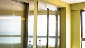 Un ascensor modern amb portes metàl·liques i panell de control, ubicat en un passadís amb terra de rajola i parets grogues.
