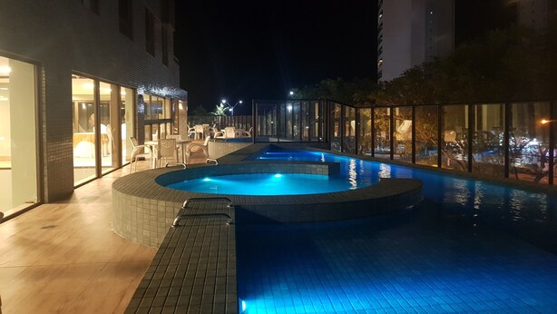 Una piscina exterior amb un solàrium de fusta, envoltada de parets de vidre i cadires, i un edifici amb una entrada il·luminada.