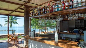 Zona de bar con vista al océano, muebles de madera y una variedad de botellas de licores.