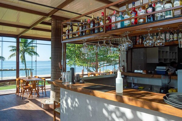 A bar area with a view of the ocean, wooden furniture, and a variety of liquor bottles. - Pousada Algodão da Praia (Araruama)