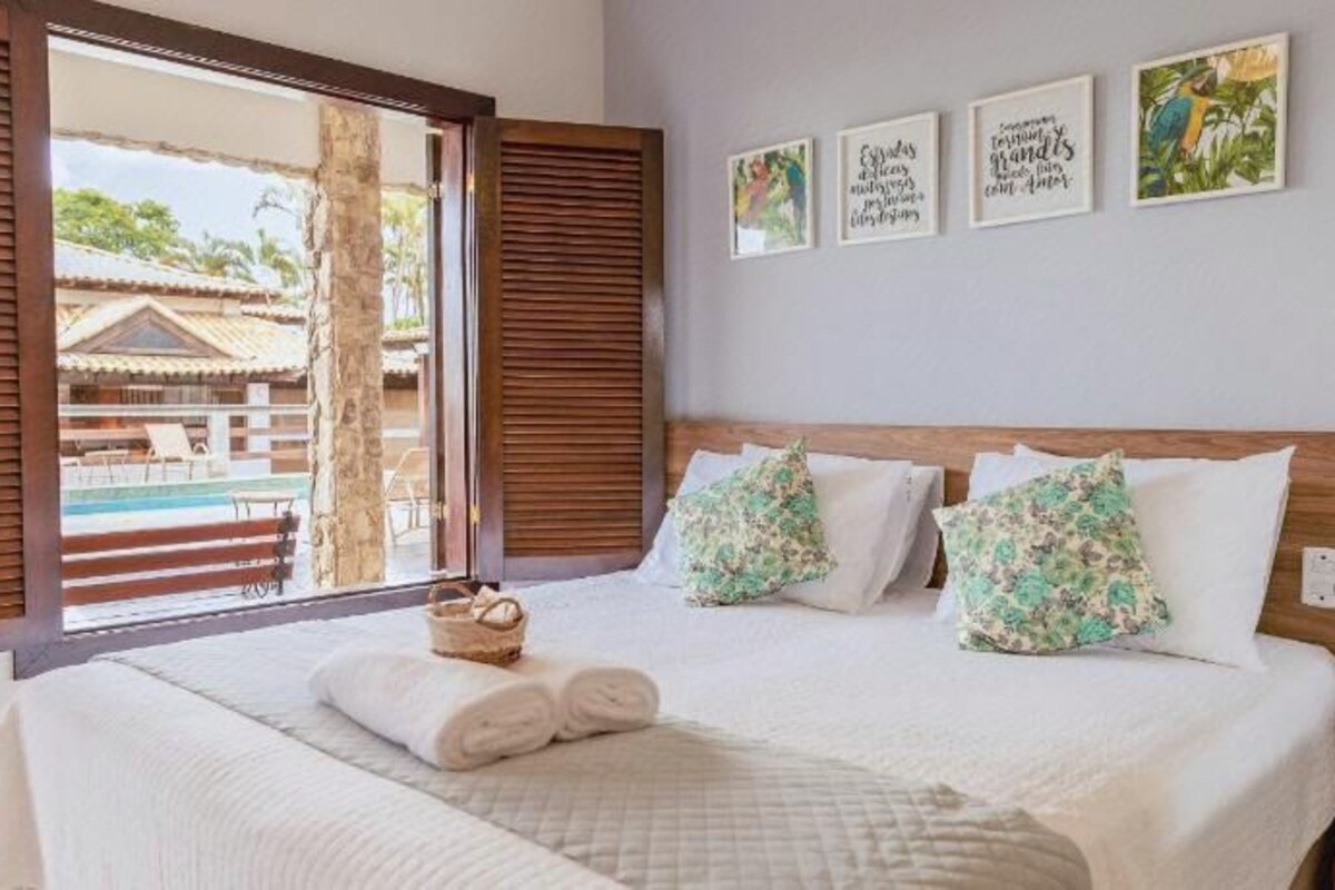 A hotel room with a bed, a nightstand, a window with wooden shutters, and a view of a pool and building outside.