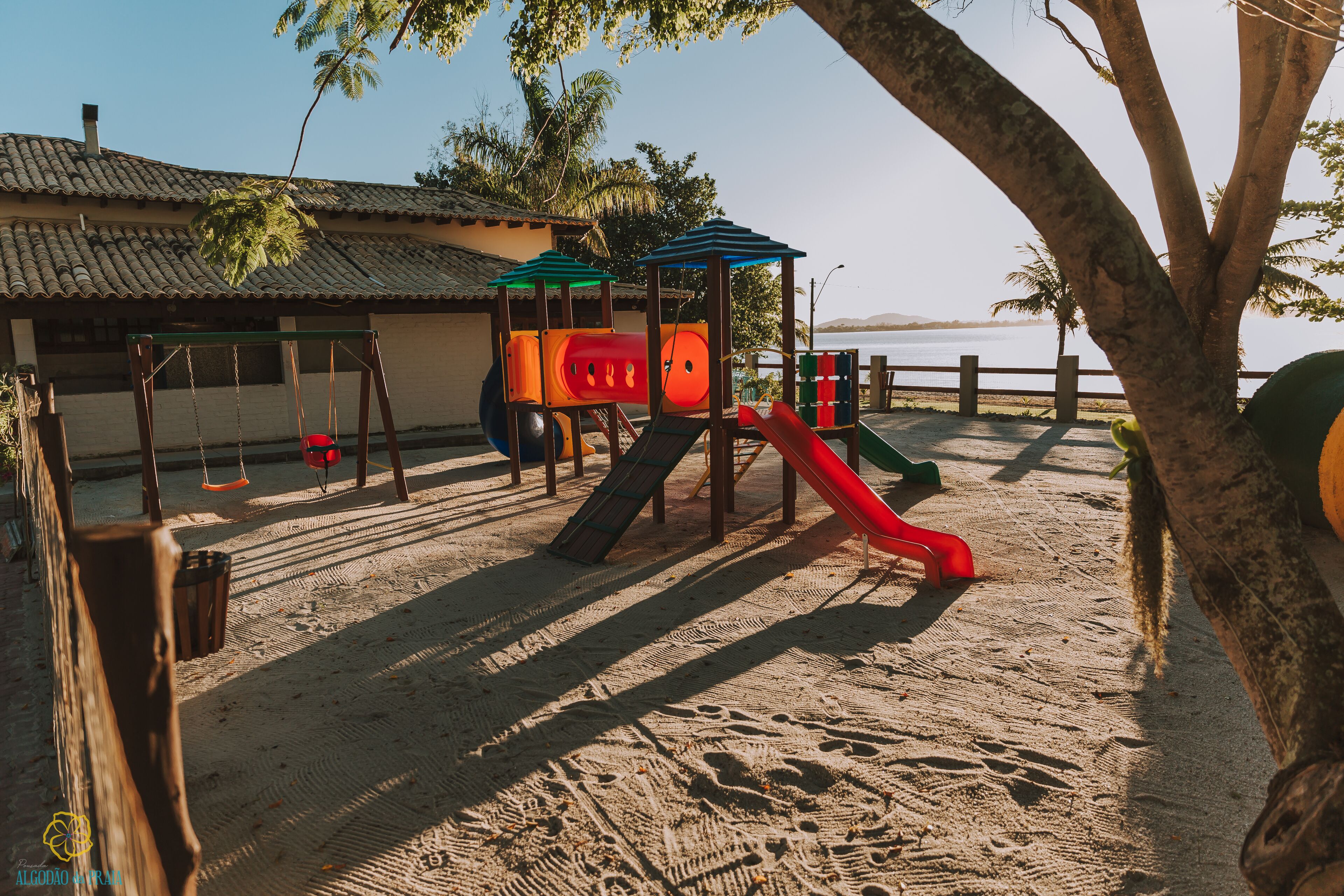 Ein Spielplatz mit Rutsche und Schaukeln, sandigem Untergrund und einem Gebäude mit Ziegeldach im Hintergrund.