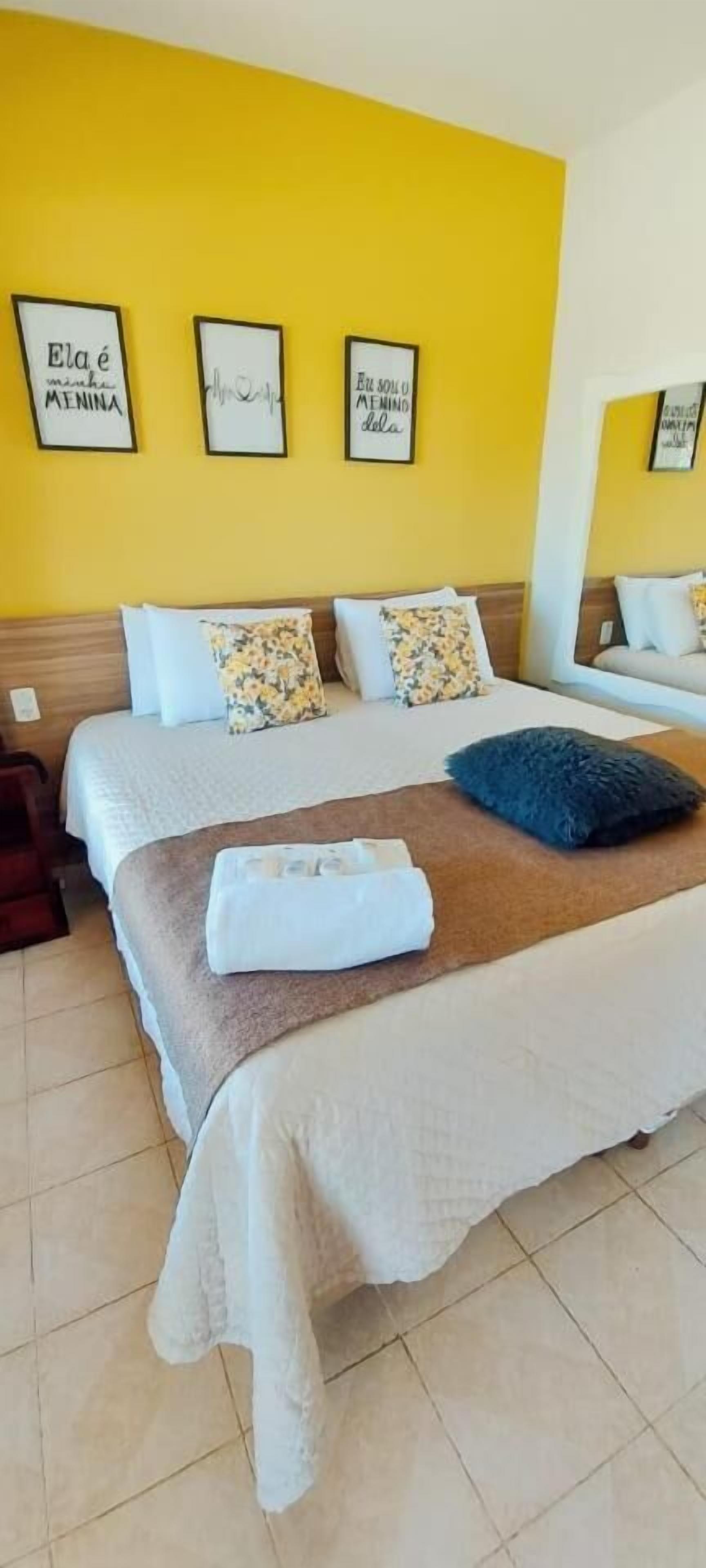 Une chambre d’hôtel avec un grand lit, deux oreillers, une couverture et une serviette pliée. Trois citations encadrées ornent le mur.