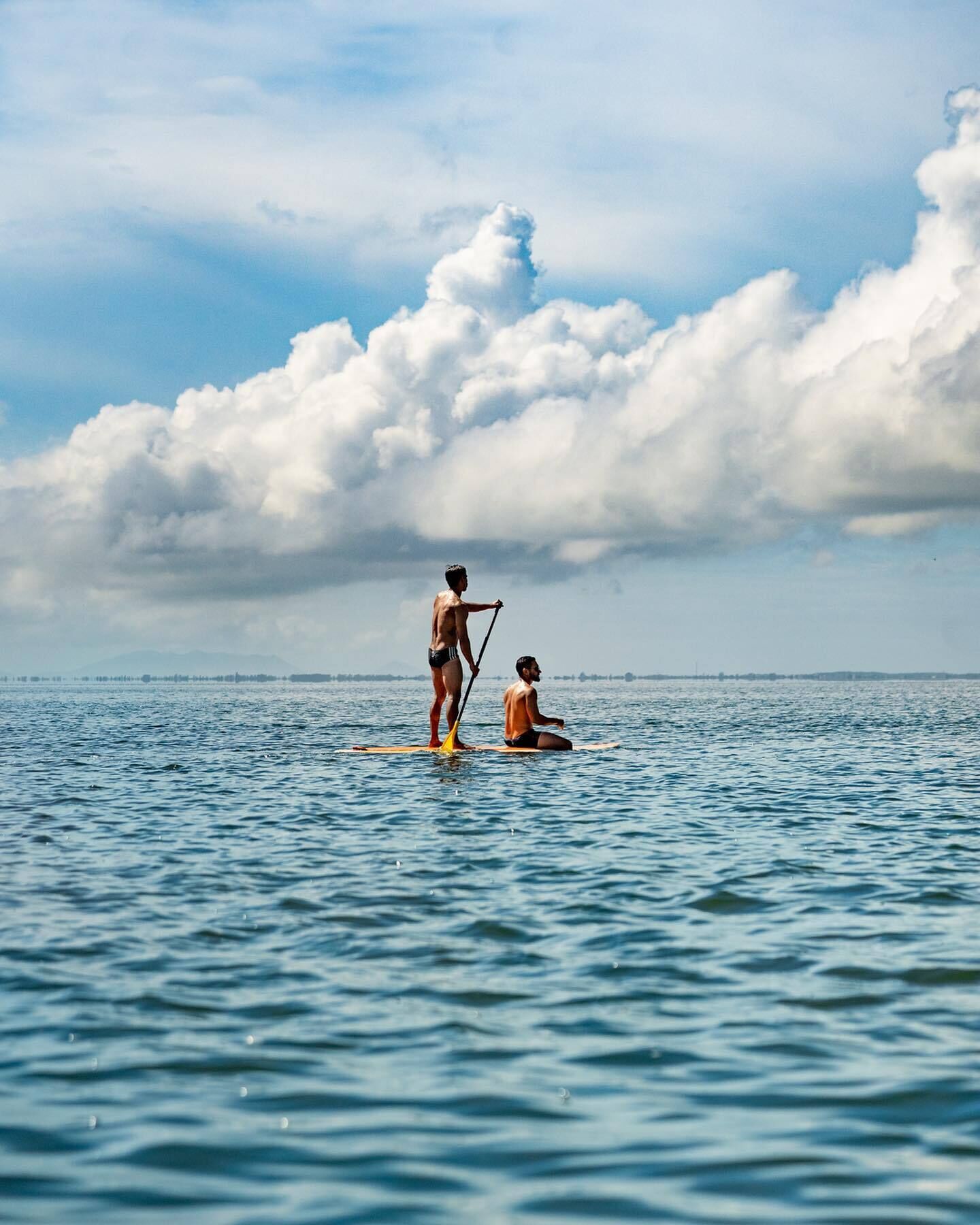 A person standing on a paddleboard in the water, another person sitting on a paddleboard, and a distant shoreline with clouds in the sky.