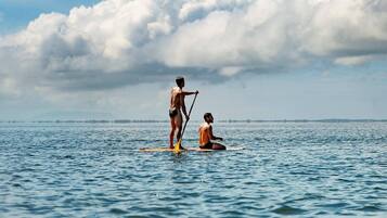 A person standing on a paddleboard in the water, another person sitting on a paddleboard, and a distant shoreline with clouds in the sky.