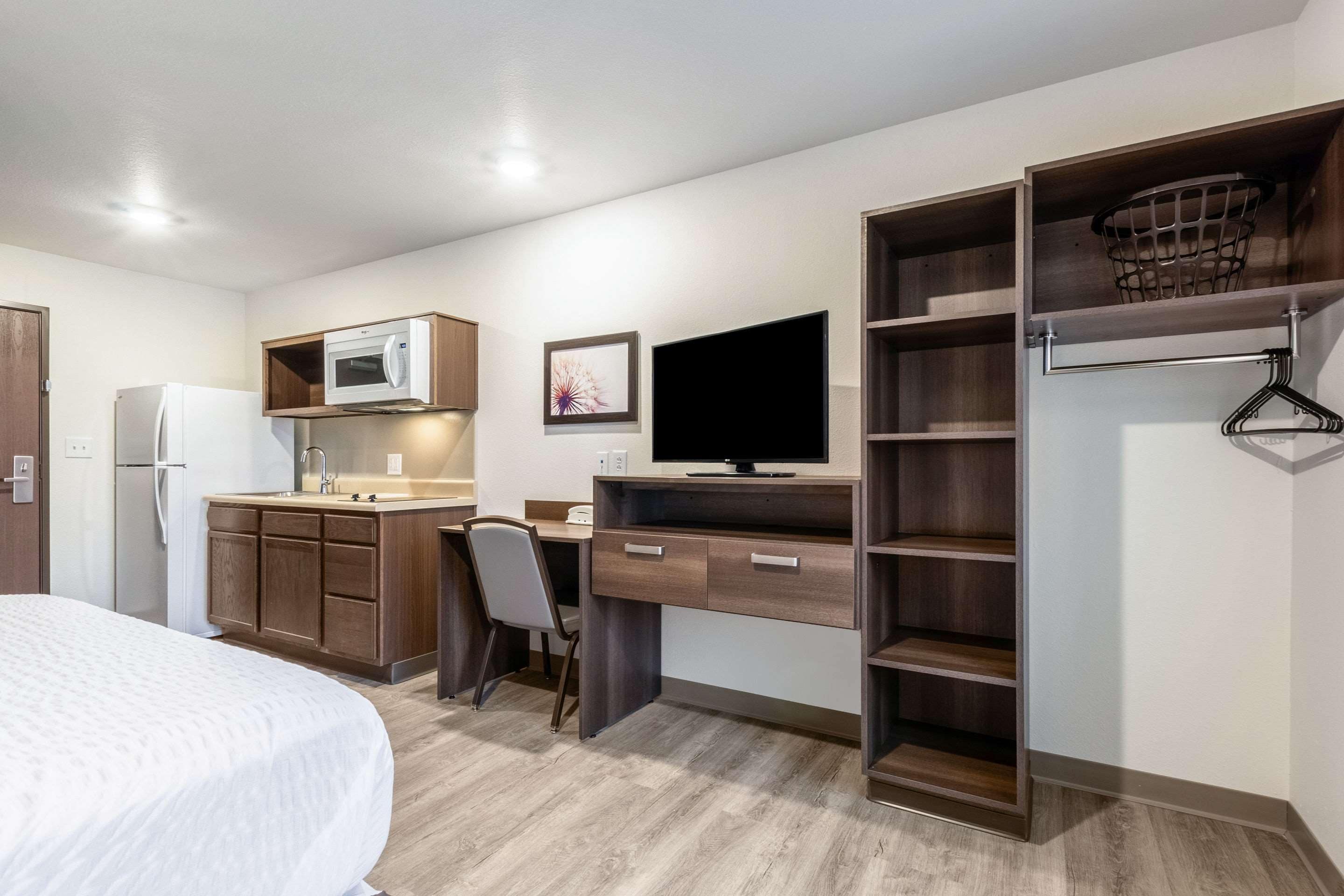 A compact hotel room with a bed, kitchenette, desk, and built-in shelving.