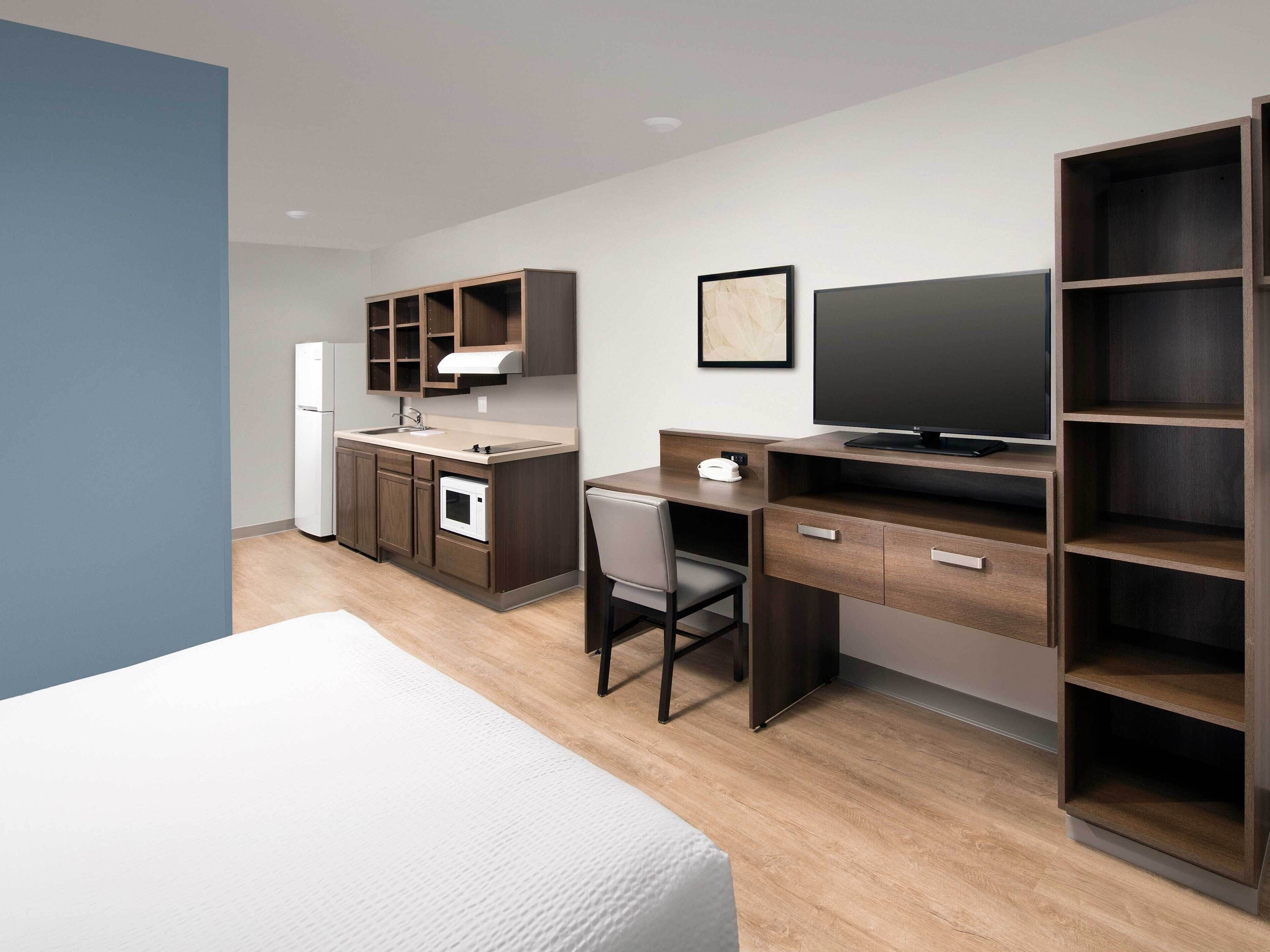 A modern hotel room with a bed, a desk, a chair, a TV, a wardrobe, a kitchen area with a fridge, microwave, and sink.