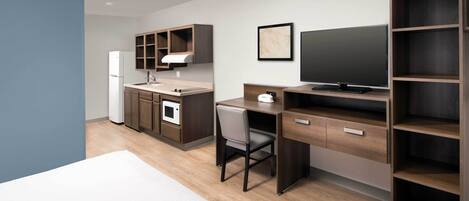 A modern hotel room with a bed, a desk, a chair, a TV, a wardrobe, a kitchen area with a fridge, microwave, and sink.