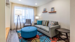 A modern living room with a grey sofa, blue ottoman, and a small round table. There are two wall-mounted shelves with pictures, a floor lamp, and a chair. - WoodSpring Suites Indianapolis Castleton (Indianapolis)